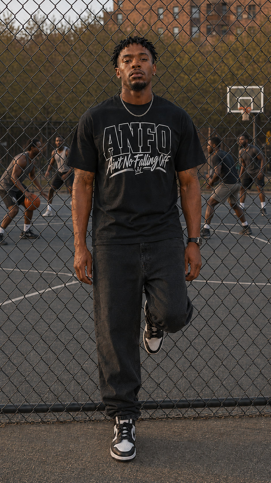 Man wearing the black ANFO Classic White Graphic Tee featuring the white “Ain’t No Falling Off” design, standing by a chain-link fence on an outdoor basketball court.