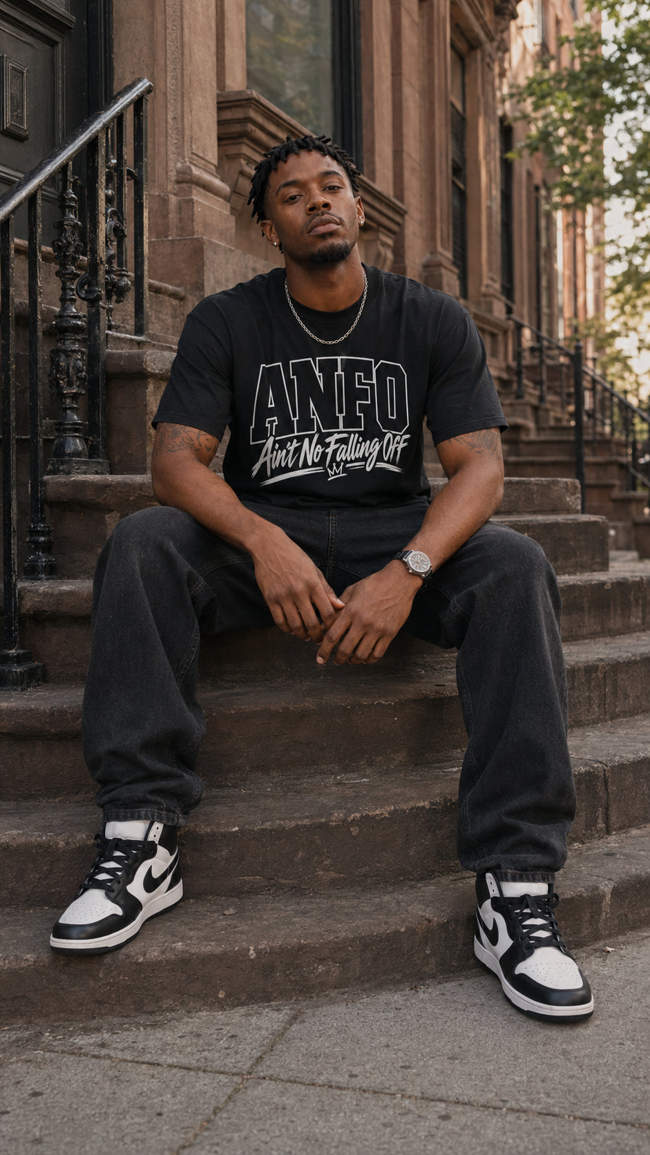 Man wearing the black ANFO Classic White Graphic Tee featuring the white “Ain’t No Falling Off” design, seated on stone steps outside a brown brick building.