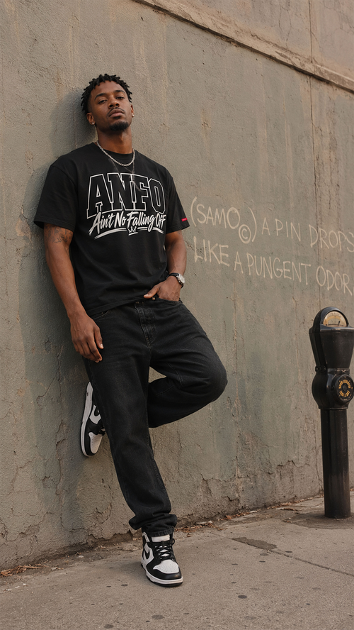 A man wearing a black graphic t-shirt with 'ANFO Ain't No Falling Off' printed on the front, black jeans, and black and white sneakers, leaning against a textured concrete wall with graffiti.