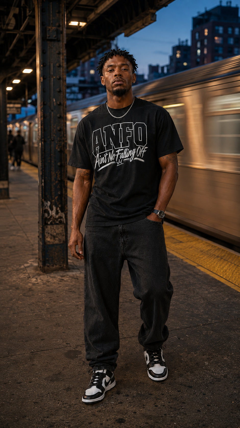 Man wearing the black ANFO Classic White Graphic Tee featuring the white “Ain’t No Falling Off” design, standing on a city train platform with a moving train in the background.