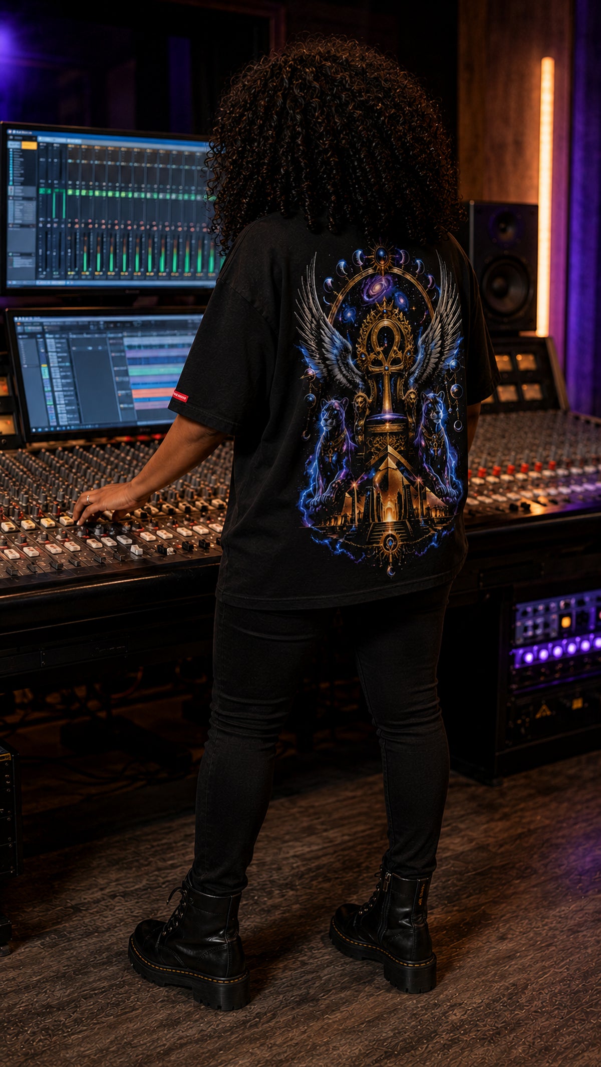 A person standing in a recording studio, viewed from behind, wearing a black graphic t-shirt with a large blue, gold, and purple winged mechanical design on the back, paired with black pants and black combat boots.