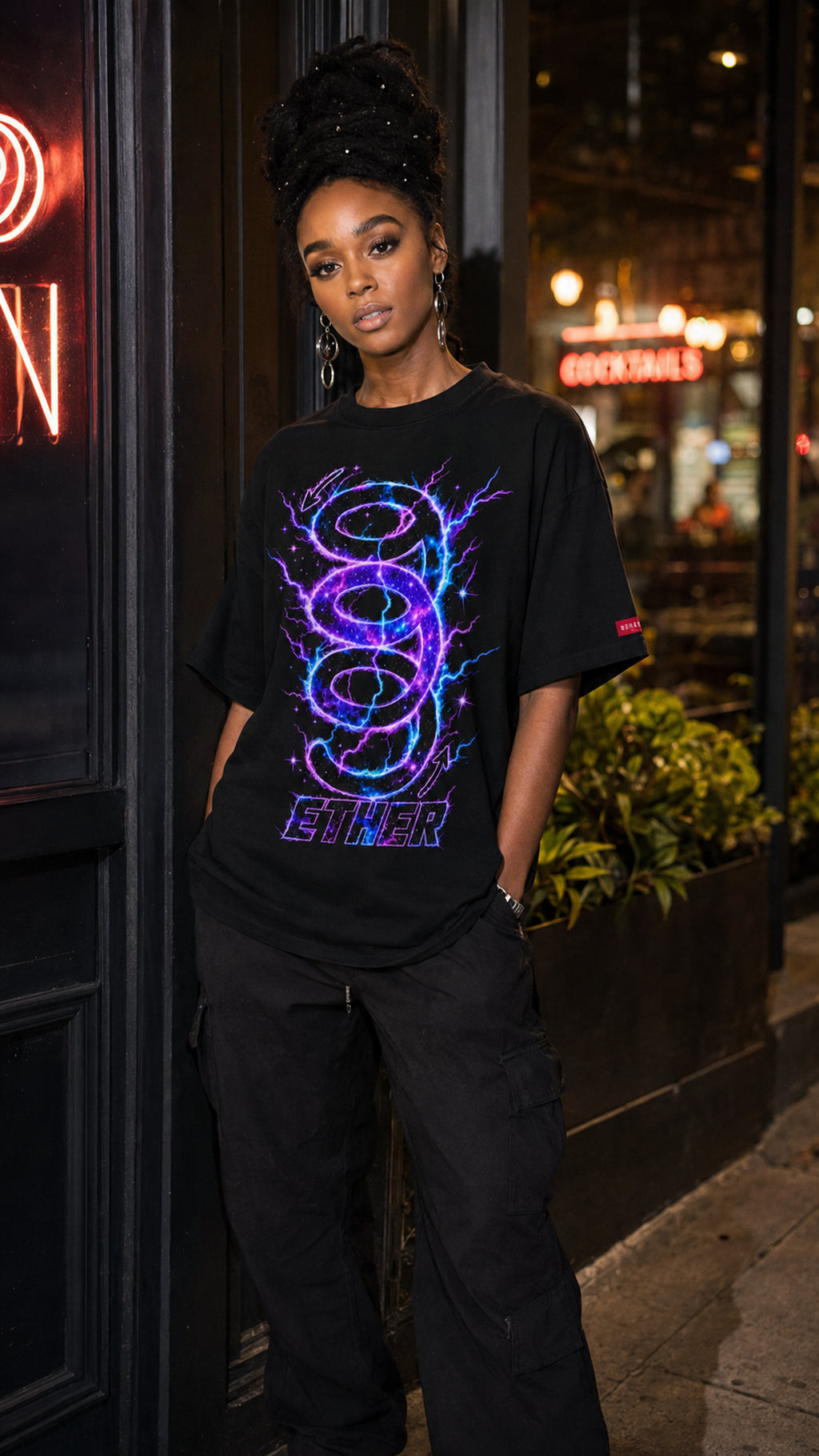 Woman wearing the black Nine (9) Ether Cosmic Energy Graphic Tee with a glowing purple-and-blue design, paired with black cargo pants outside a restaurant at night.