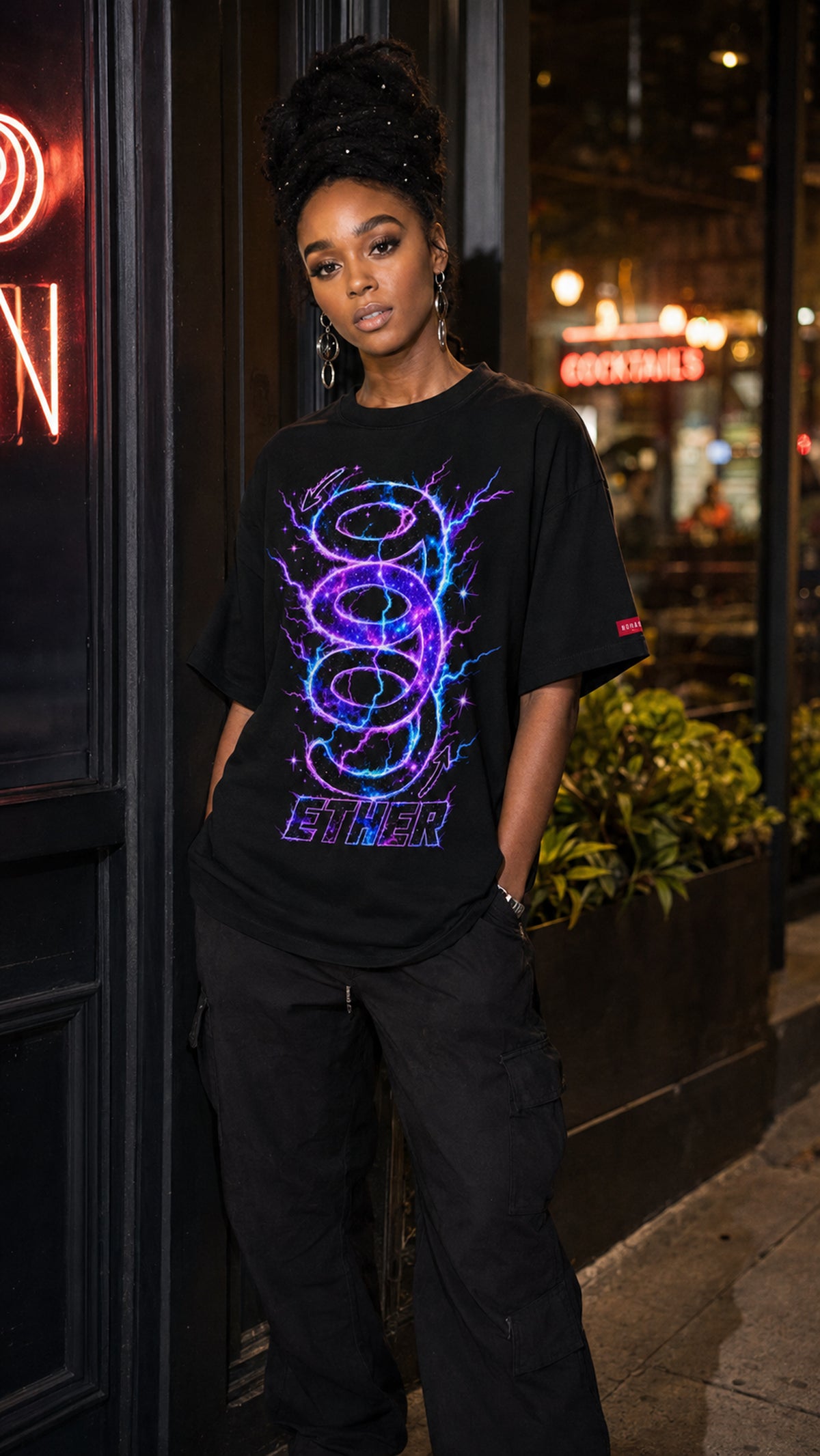 Woman wearing the black Nine (9) Ether Cosmic Energy Graphic Tee with a glowing purple-and-blue design, paired with black cargo pants outside a restaurant at night.