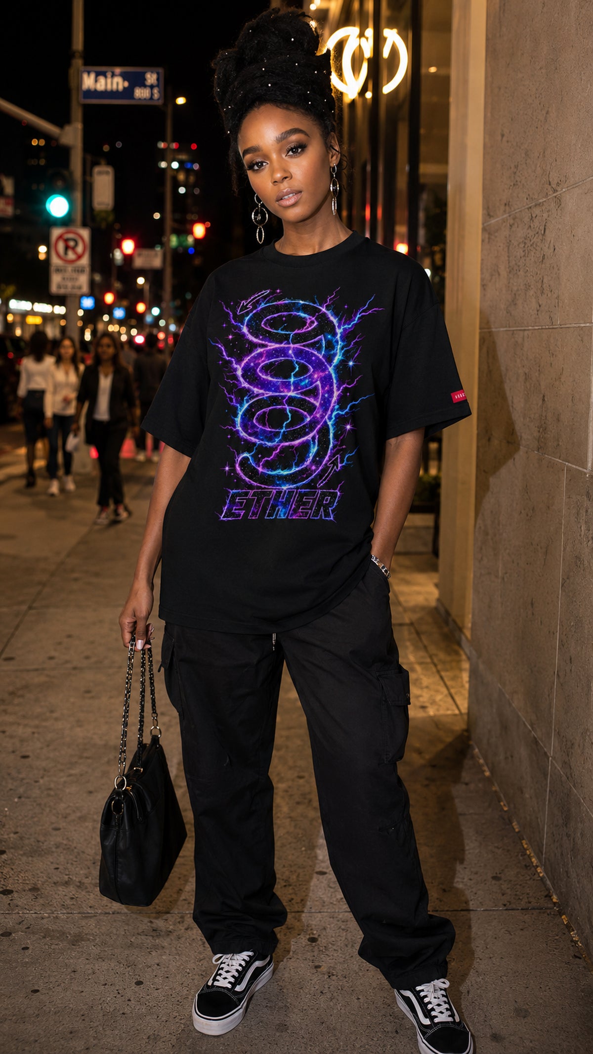Woman wearing the black Nine (9) Ether Cosmic Energy Graphic Tee with a glowing purple-and-blue design, black cargo pants and sneakers, holding a handbag on a city sidewalk at night.