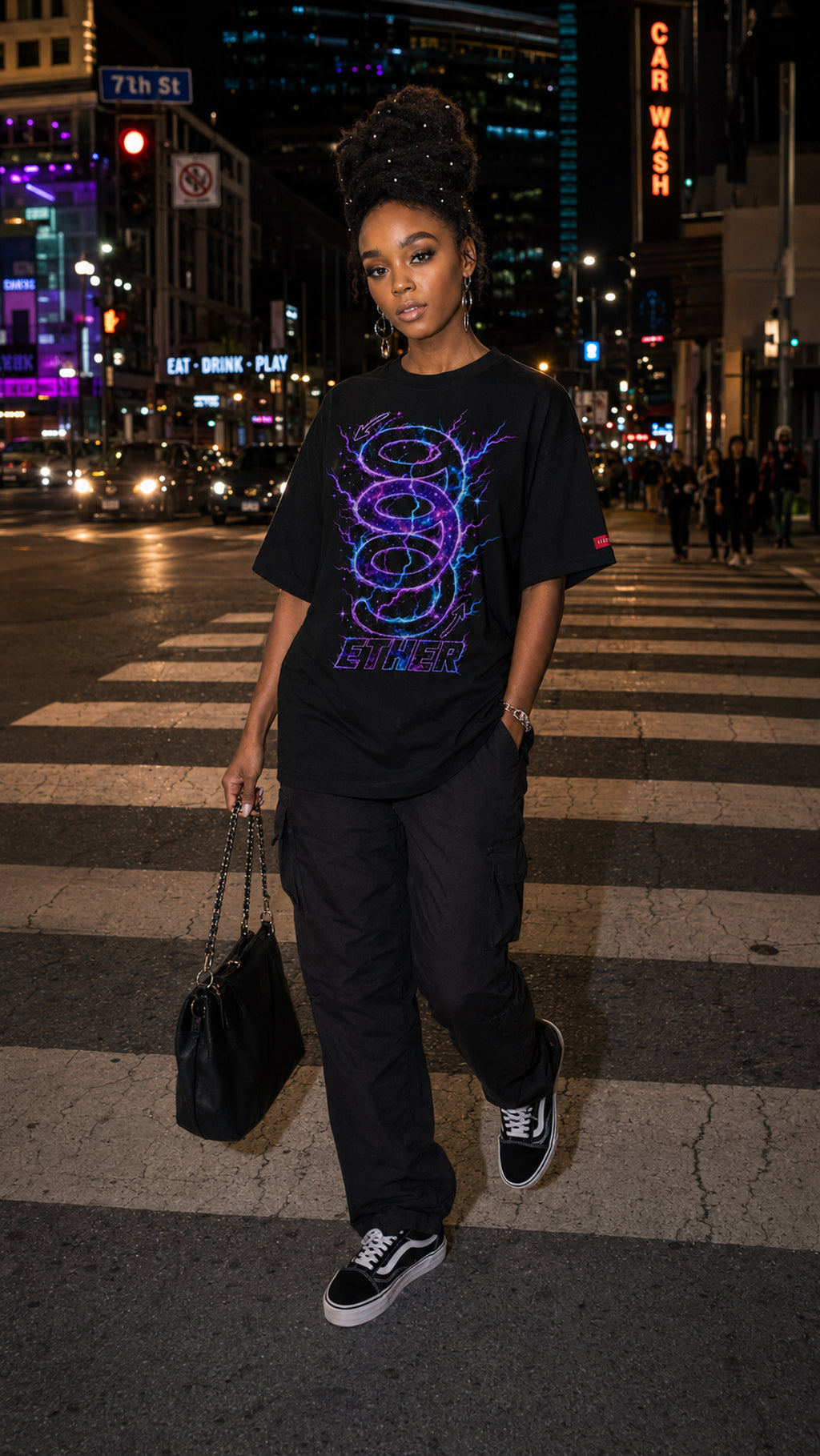 Woman wearing the oversized black Nine (9) Ether Cosmic Energy Graphic Tee with a glowing purple-and-blue design, crossing a city street at night with neon signs in the background.