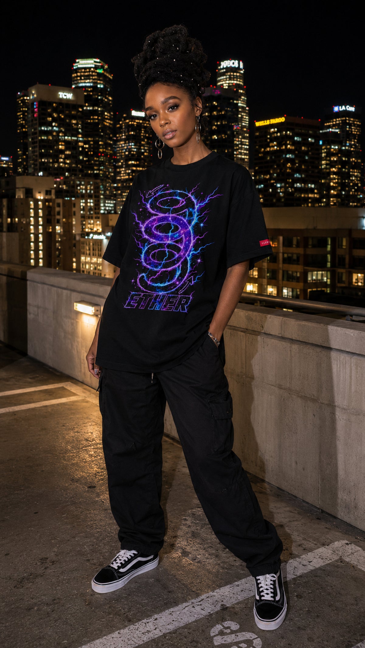 Model wearing the black Nine (9) Ether Cosmic Energy Graphic Tee with a glowing purple-and-blue design, black cargo pants and sneakers, on a rooftop overlooking the city at night.